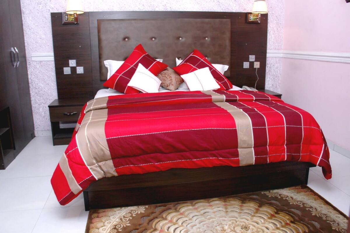 A neatly made bed with a red and beige bedspread and matching pillows, a wooden headboard, and bedside lamps.