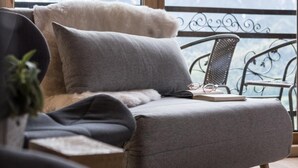 Un coin salon confortable avec un canapé et un fauteuil, une petite table avec un livre et des lunettes, et un balcon doté d’une rambarde en métal et offrant une vue sur les montagnes.