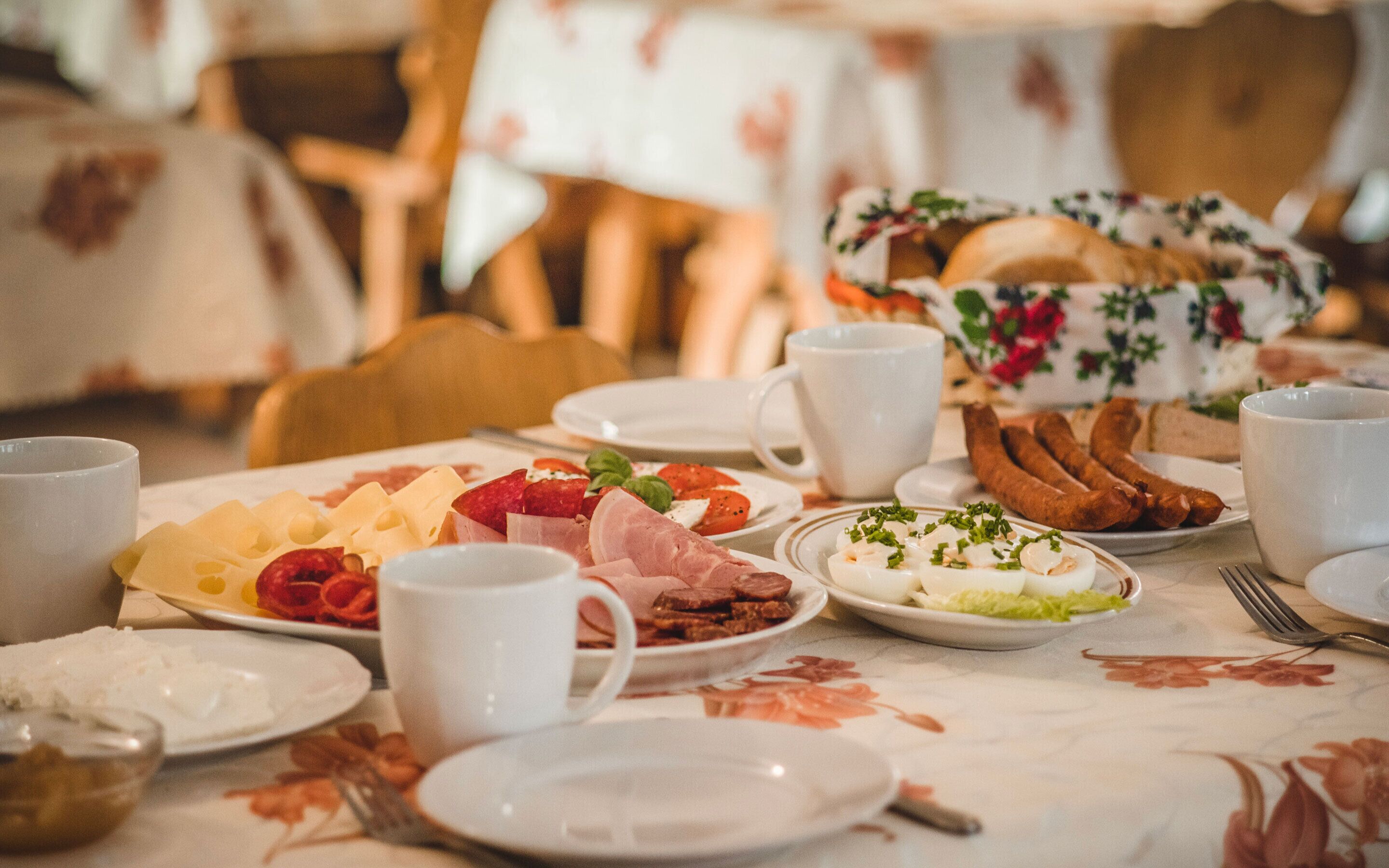 Une table mise pour le petit-déjeuner, avec des assiettes remplies de nourriture, notamment du fromage, de la charcuterie et des œufs, ainsi que plusieurs tasses blanches.