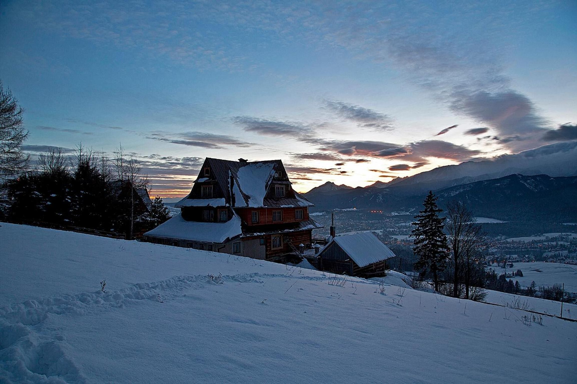 Una casa de madera de dos pisos con techo nevado, ubicada frente a un paisaje montañoso durante el atardecer.