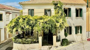 A two-story building with a tiled roof, green shutters, and a garden area with flowering plants.