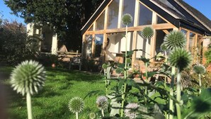 Une maison traditionnelle avec un toit en pente et de grandes fenêtres, entourée d’un jardin bien entretenu où poussent différentes espèces de plantes.