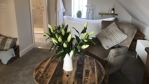 Un salon chaleureux avec une table basse ronde en bois, un fauteuil beige et un vase blanc contenant des plantes vertes. On aperçoit une grande baignoire au fond, et une fenêtre laisse entrer la lumière naturelle.