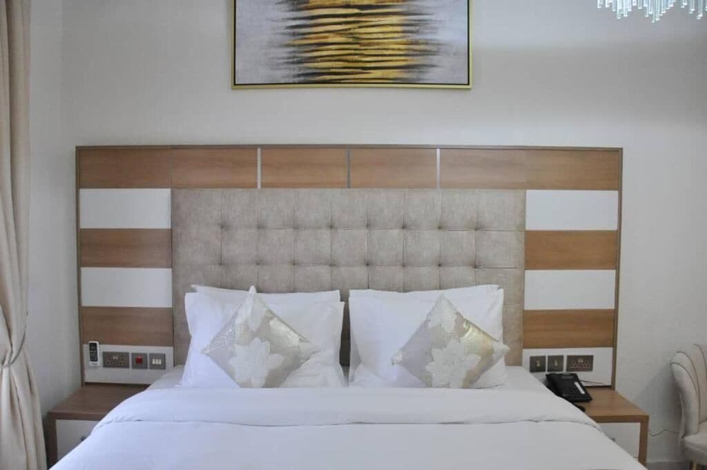 Une chambre d’hôtel avec une tête de lit capitonnée, un lit avec une literie blanche et des coussins décoratifs, une table de chevet avec un téléphone et une œuvre d’art accrochée au mur.