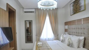 A hotel room with a bed, a television, a chandelier, and a large window with curtains. - Brighams Suites (Lagos)