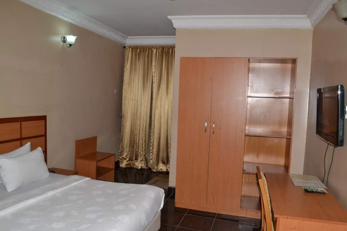 Habitación de hotel con una cama, un armario de madera, un escritorio con una silla, un televisor y una cortina.