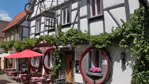 A traditional half-timbered building with a red tiled roof, outdoor seating with red umbrellas, and a sign indicating a summer bowtie event. - homie hotel oechsle & brix (Sommerhausen)