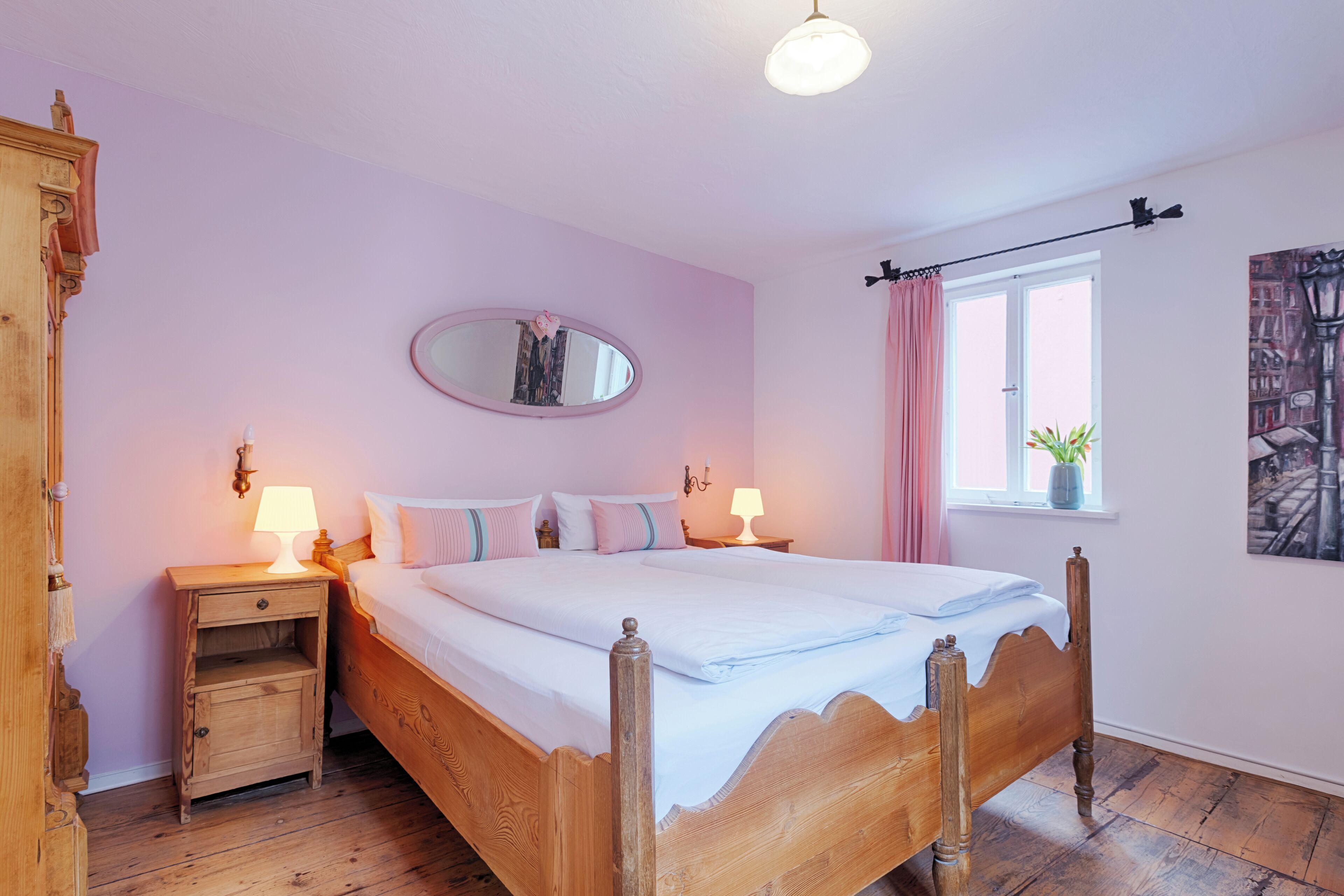 A bedroom with a wooden bed, two bedside tables with lamps, a mirror, a window with curtains, and a plant on the windowsill.
