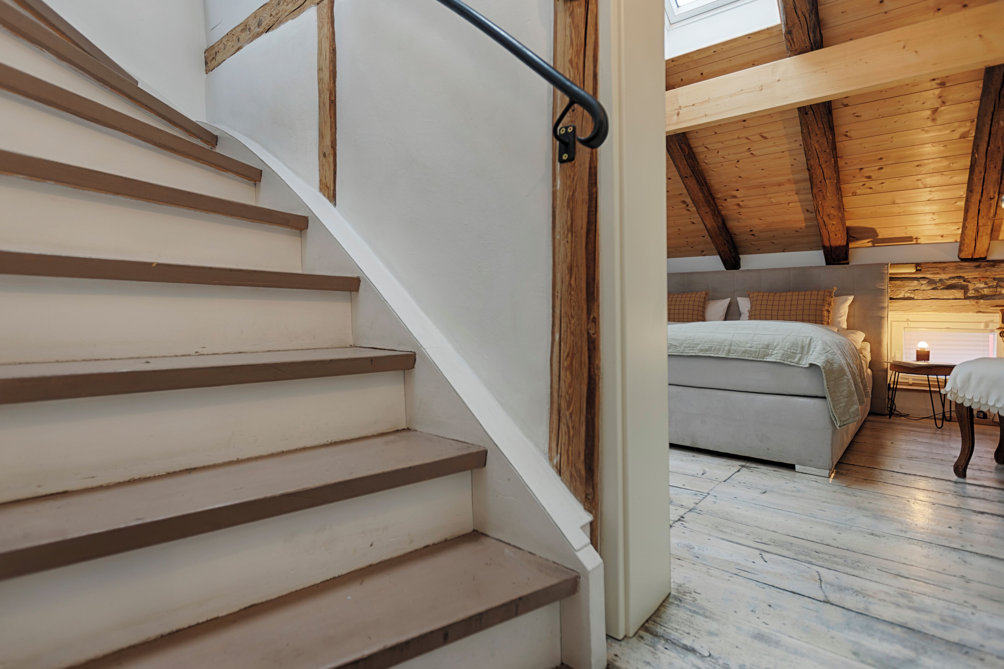 Una escalera de madera con barandilla metálica que conduce a un dormitorio con cama y mesita de noche.