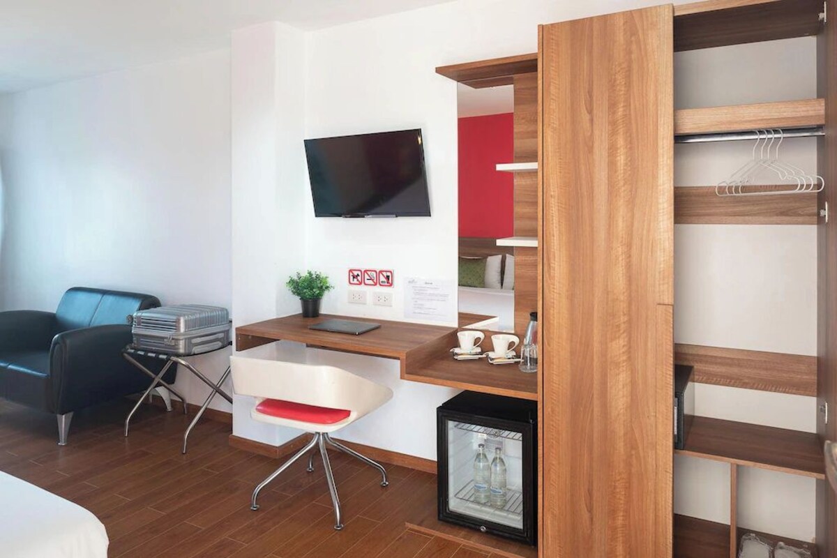 Une chambre d’hôtel comprenant un lit, un bureau avec une chaise, une télévision, un petit réfrigérateur et une armoire.