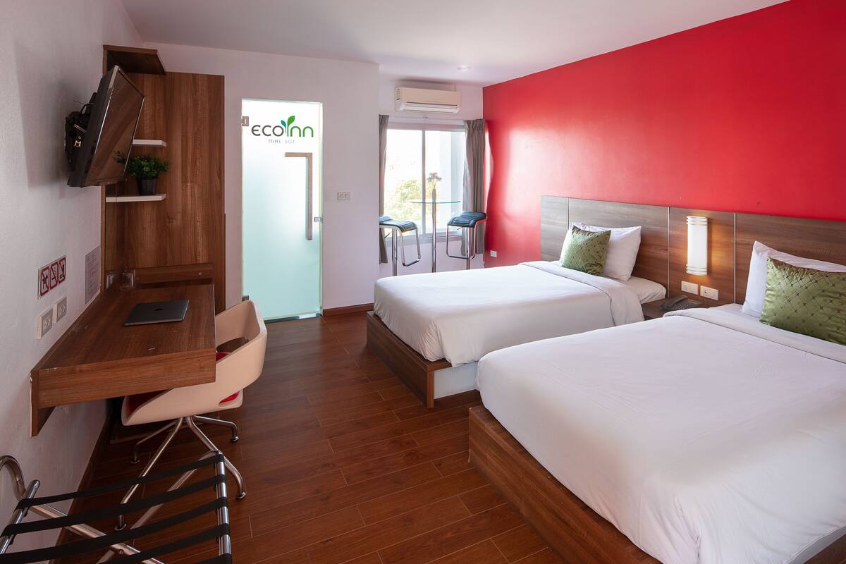 Une chambre d’hôtel avec deux lits, un bureau avec un ordinateur portable, une chaise et un mur d’accent rouge.