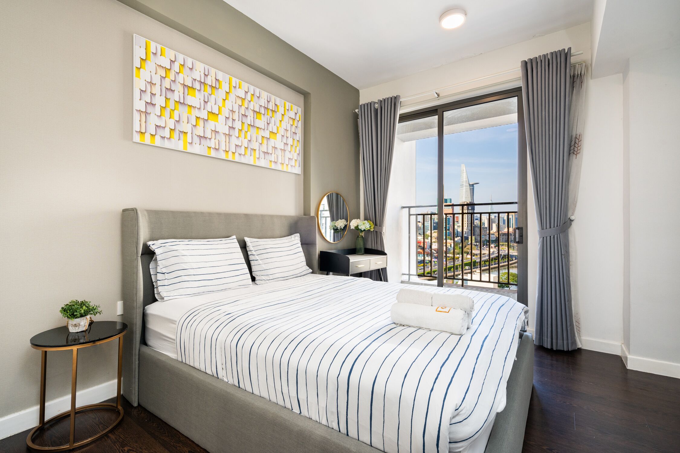 Ein modernes Schlafzimmer mit einem großen Bett, einem kleinen Nachttisch, einer Wanddekoration und Blick auf eine Stadtlandschaft durch einen Balkon.