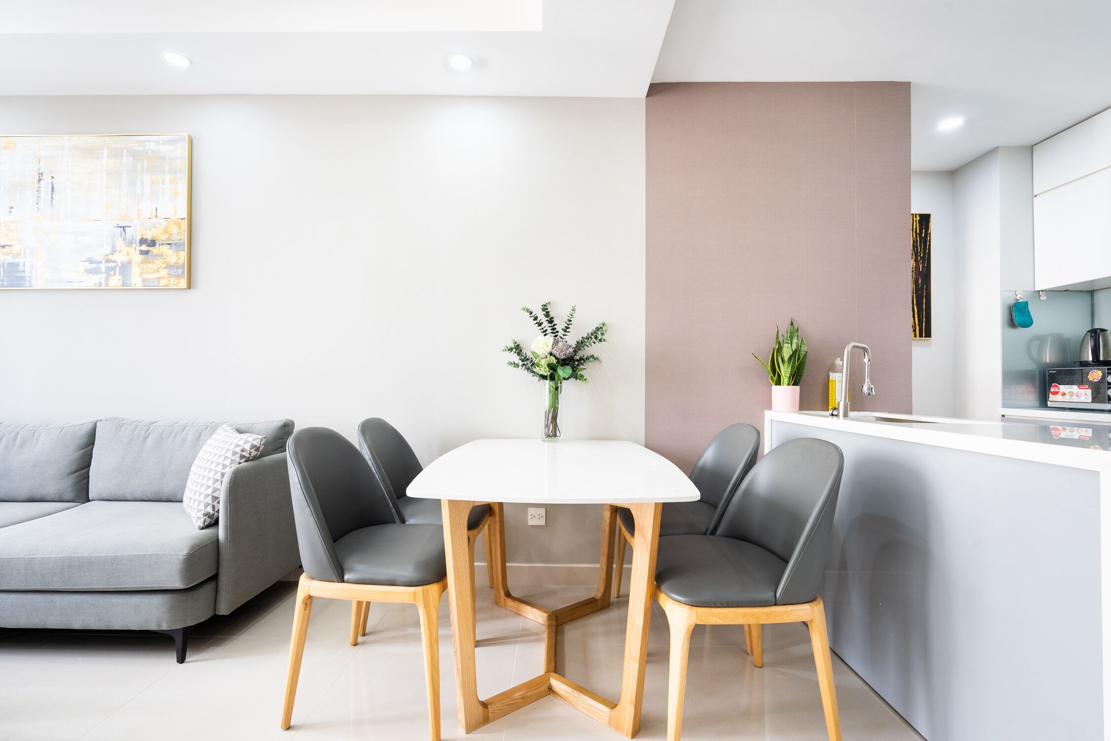 A modern living room with a grey sofa, a white dining table with four chairs, a vase with greenery, and a kitchen area with cabinets and appliances.