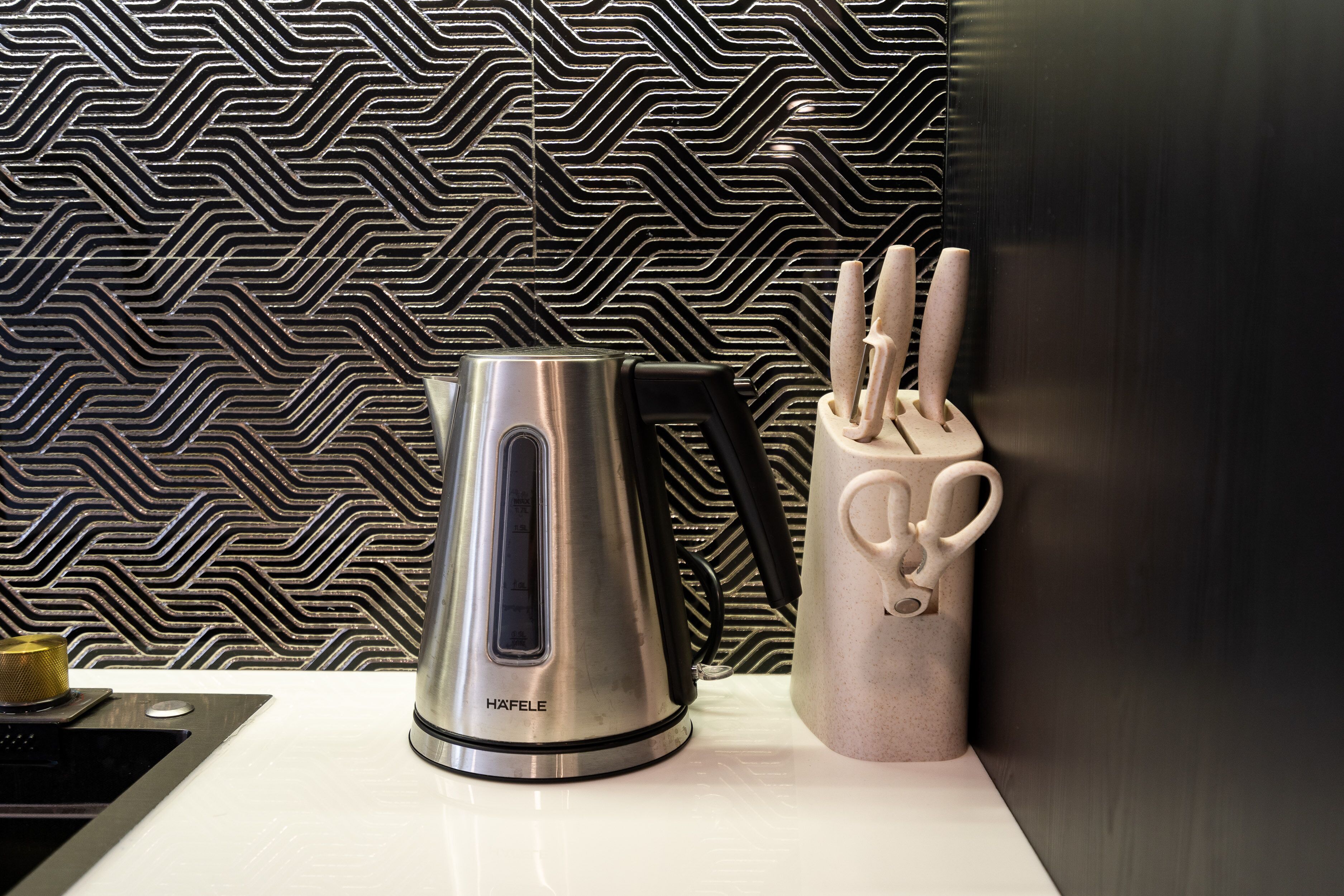 A kitchen with a modern design, featuring a stainless steel kettle, a knife block, and a patterned backsplash.