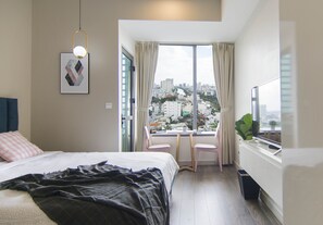 A bedroom with a bed, a desk, two chairs, a window with a city view, and a plant on the windowsill. - SAIGON 9 - Rivergate Residence Infinity Pool (Ho Chi Minh City)