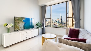 Een moderne woonkamer met een groot raam, een flatscreen televisie, een witte console, een ronde tafel en een bank met kussens.
