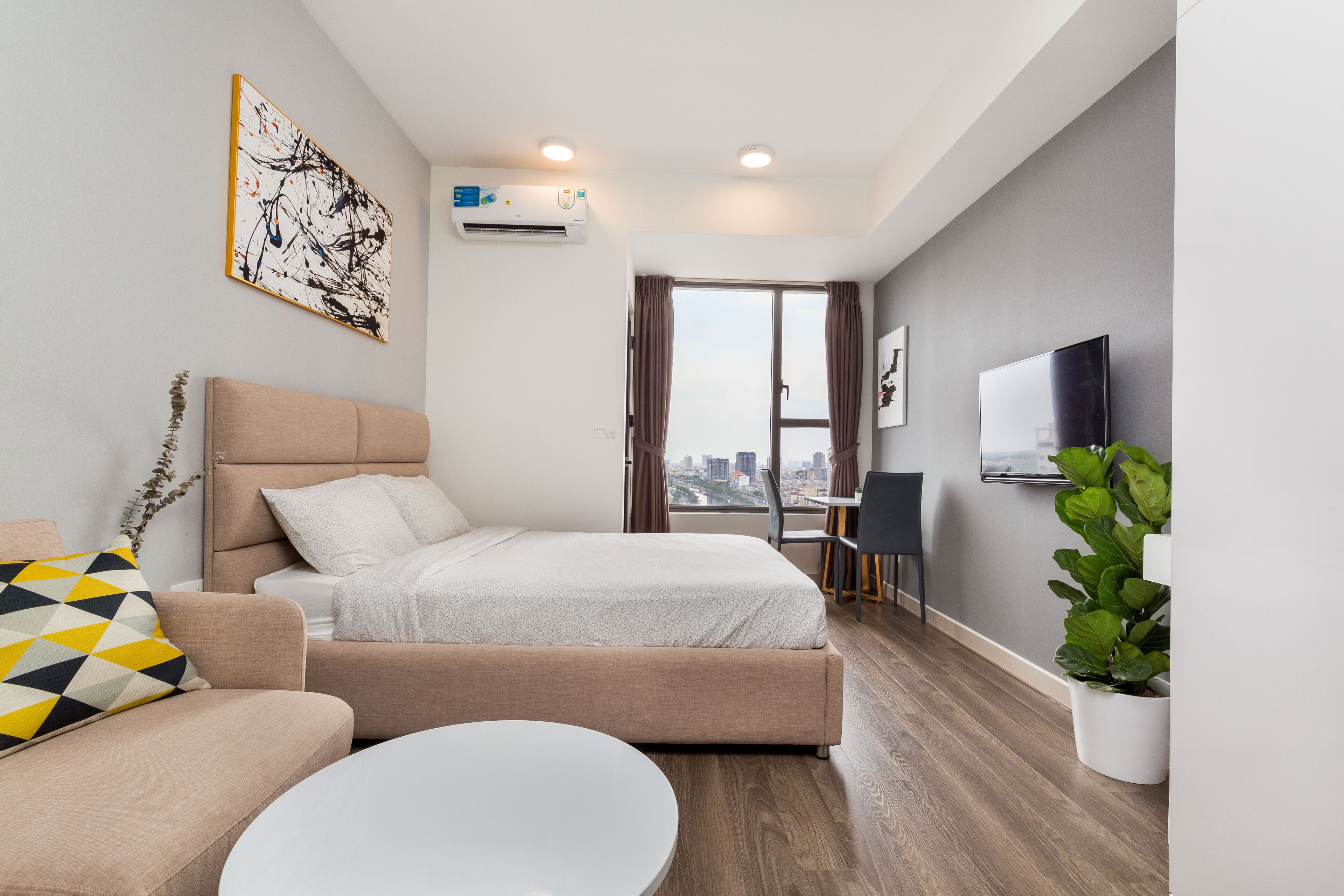 A modern hotel room with a bed, a sofa, a dining table, a potted plant, and a city view.