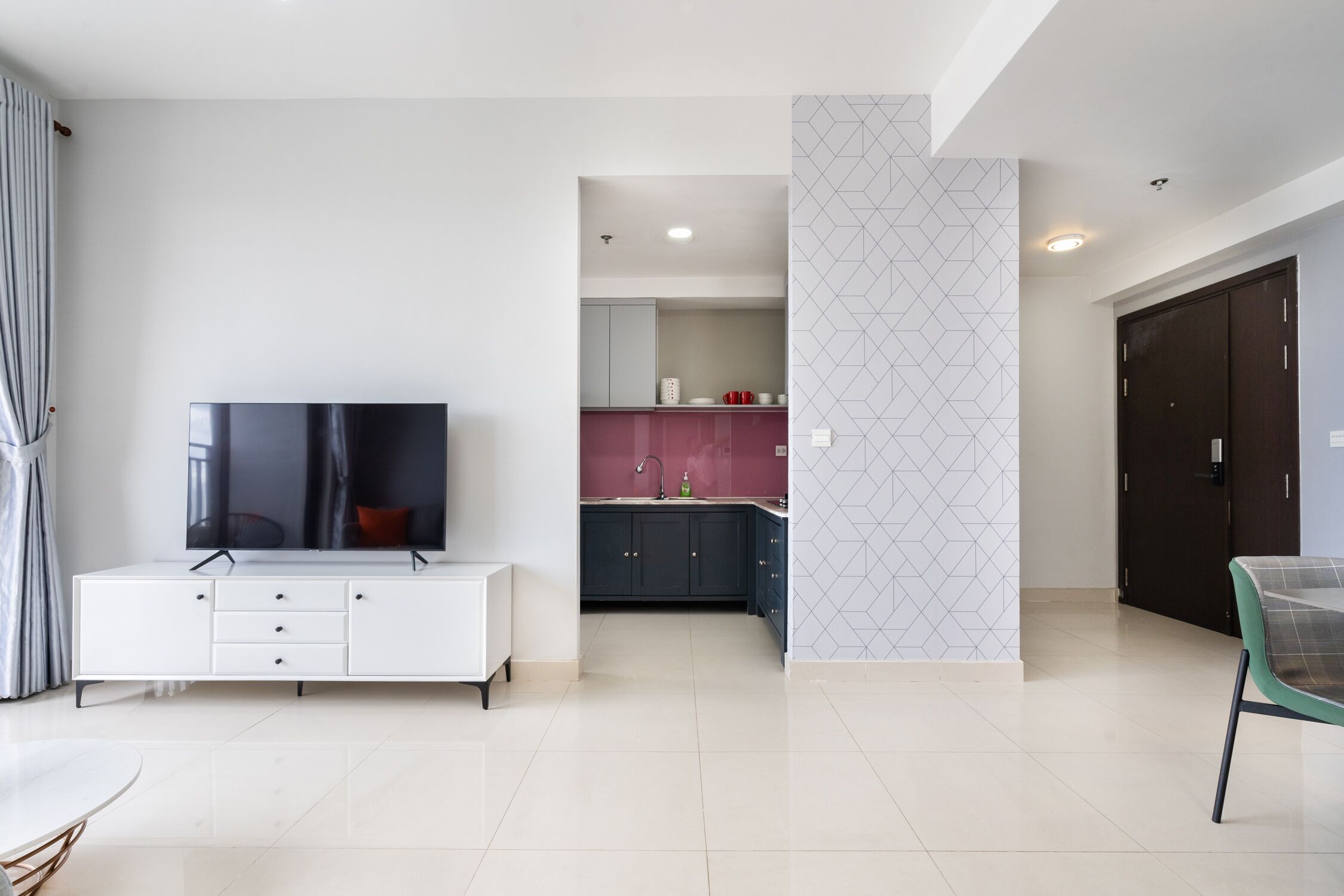 Una sala de estar moderna con un televisor de pantalla plana sobre un mueble blanco, una mesa de comedor y una cocina al fondo.