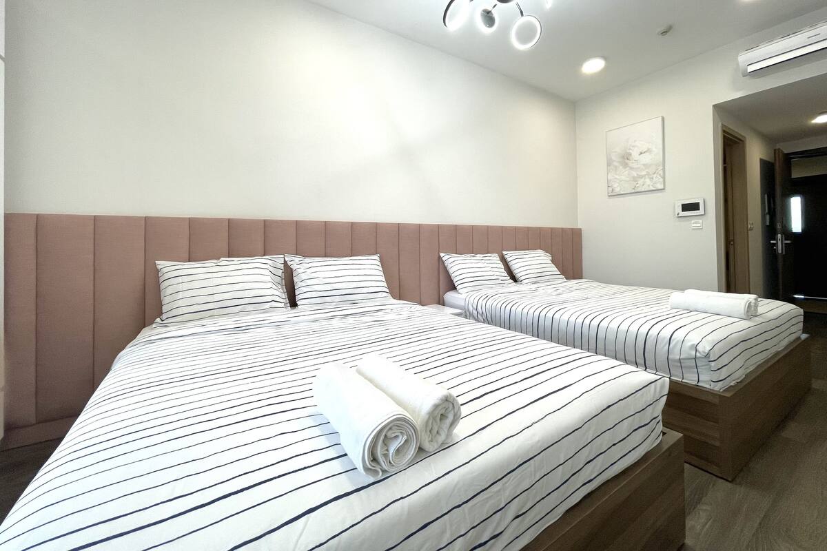 A hotel room with two beds, a modern ceiling light fixture, an air conditioning unit, and a door leading to another room.