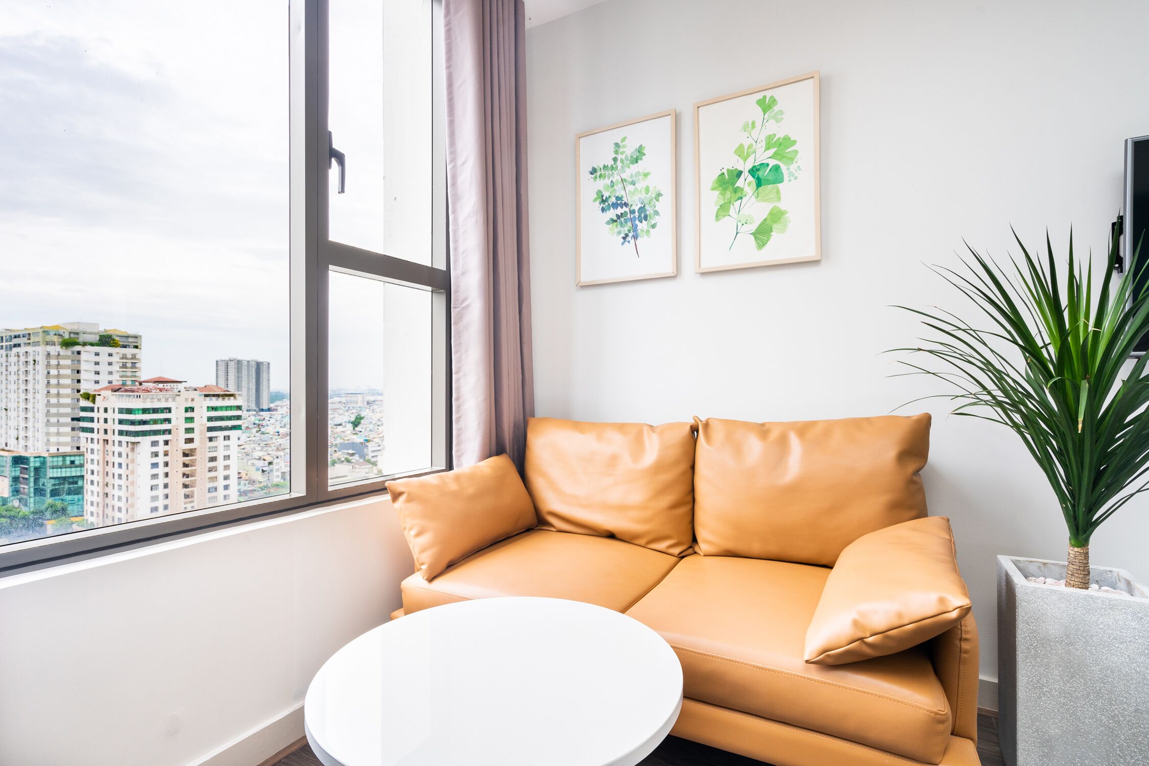 Una sala de estar moderna con un sofá de cuero color café claro, una mesa redonda blanca y un gran ventanal con vistas a la ciudad.
