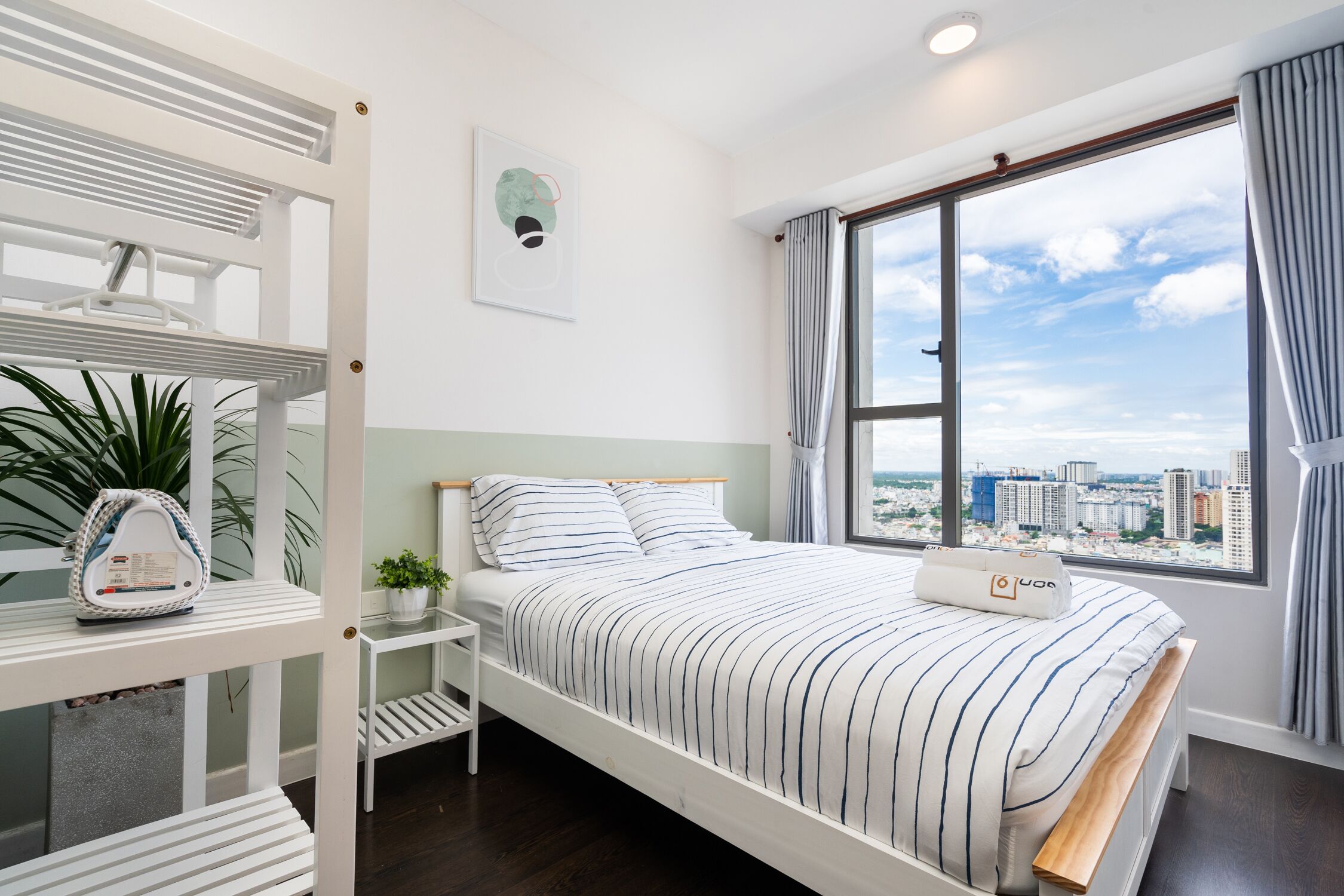 A modern bedroom with a large window, a bed with striped bedding, a small bedside table with a plant, and a white radiator.