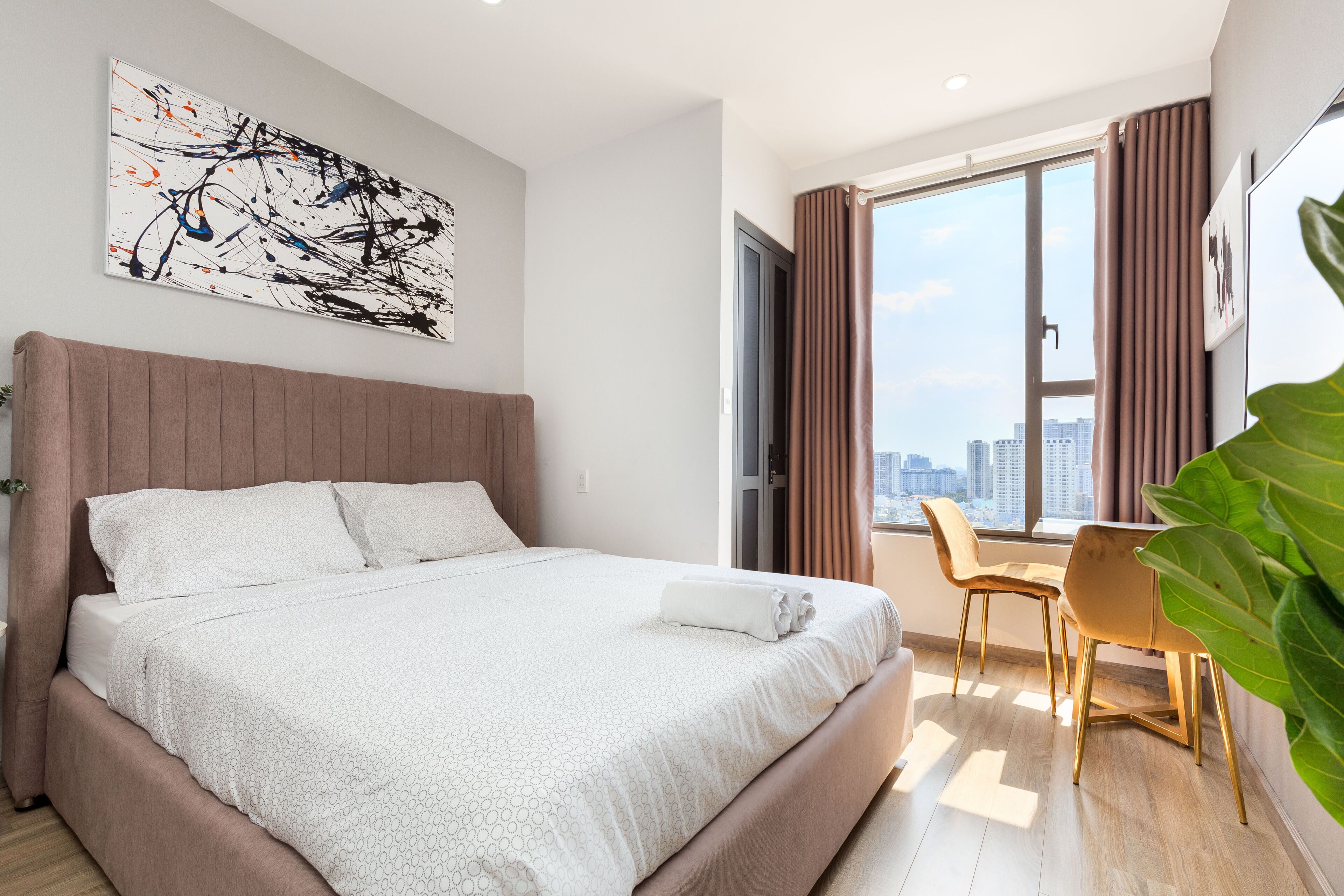 A modern hotel room with a large bed, a city view, a dining area, and a large plant.