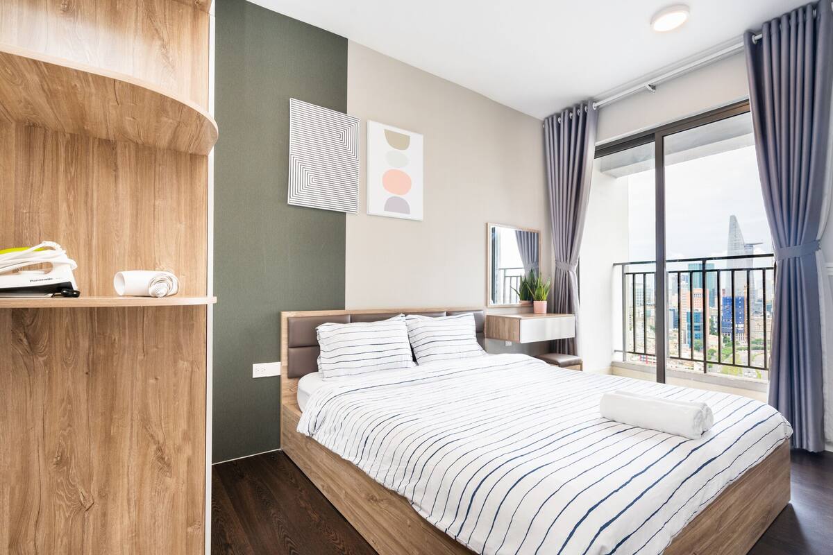 A modern hotel room with a wooden bed frame, a large window with curtains, a small bedside table with a plant, and a wall-mounted artwork.