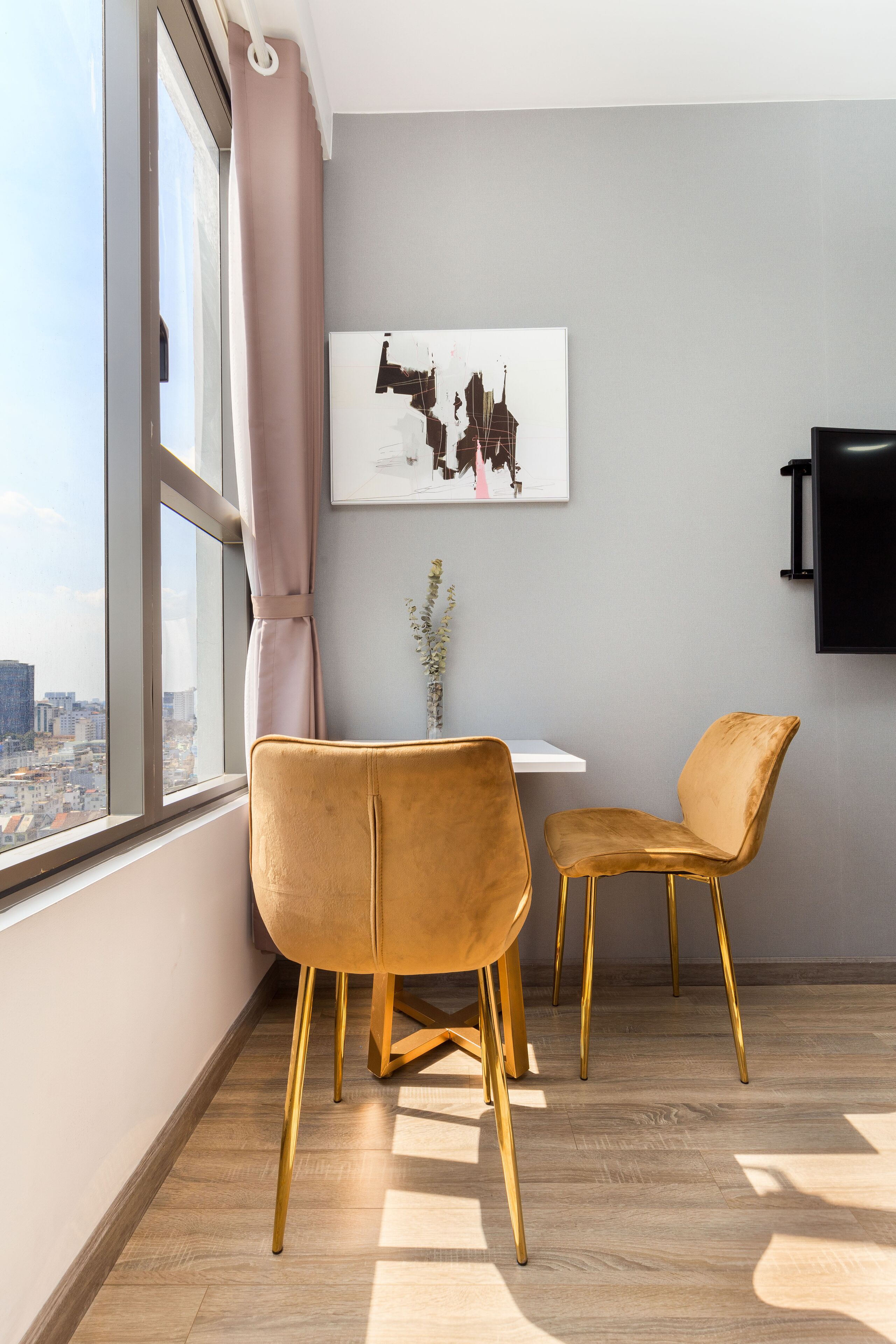 Una habitación moderna con un ventanal grande, dos sillas doradas, una mesita y un televisor montado.
