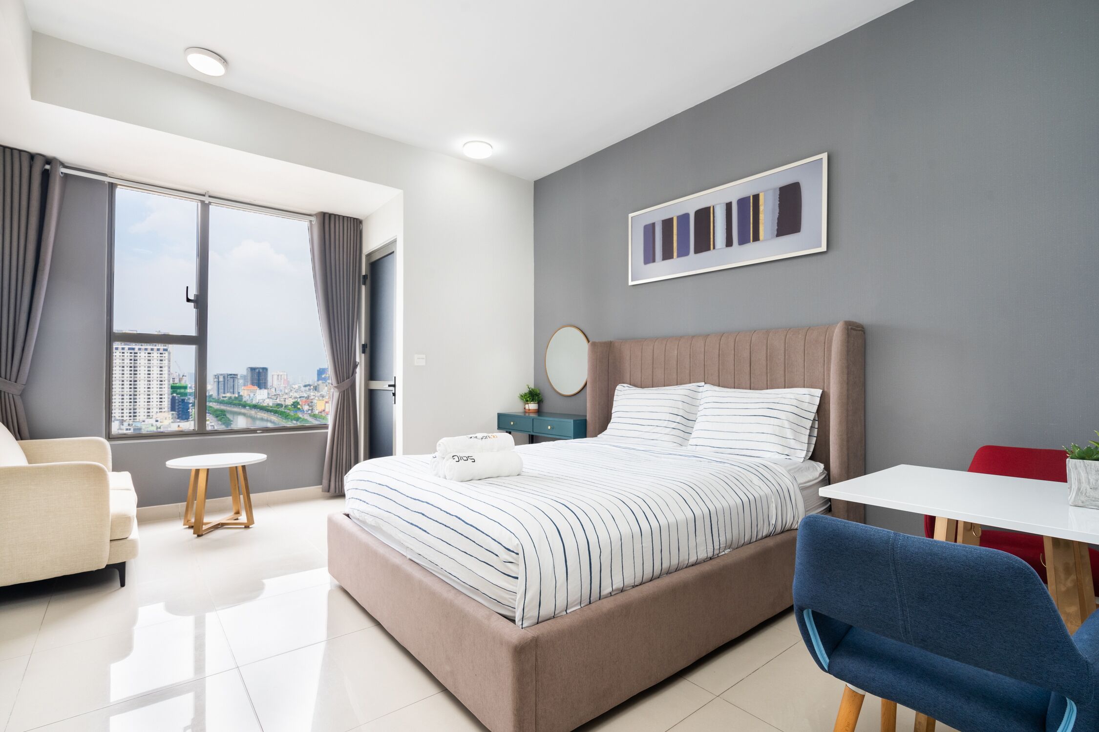 A modern hotel room with a large bed, a small table, a chair, and a view of the city through the window.