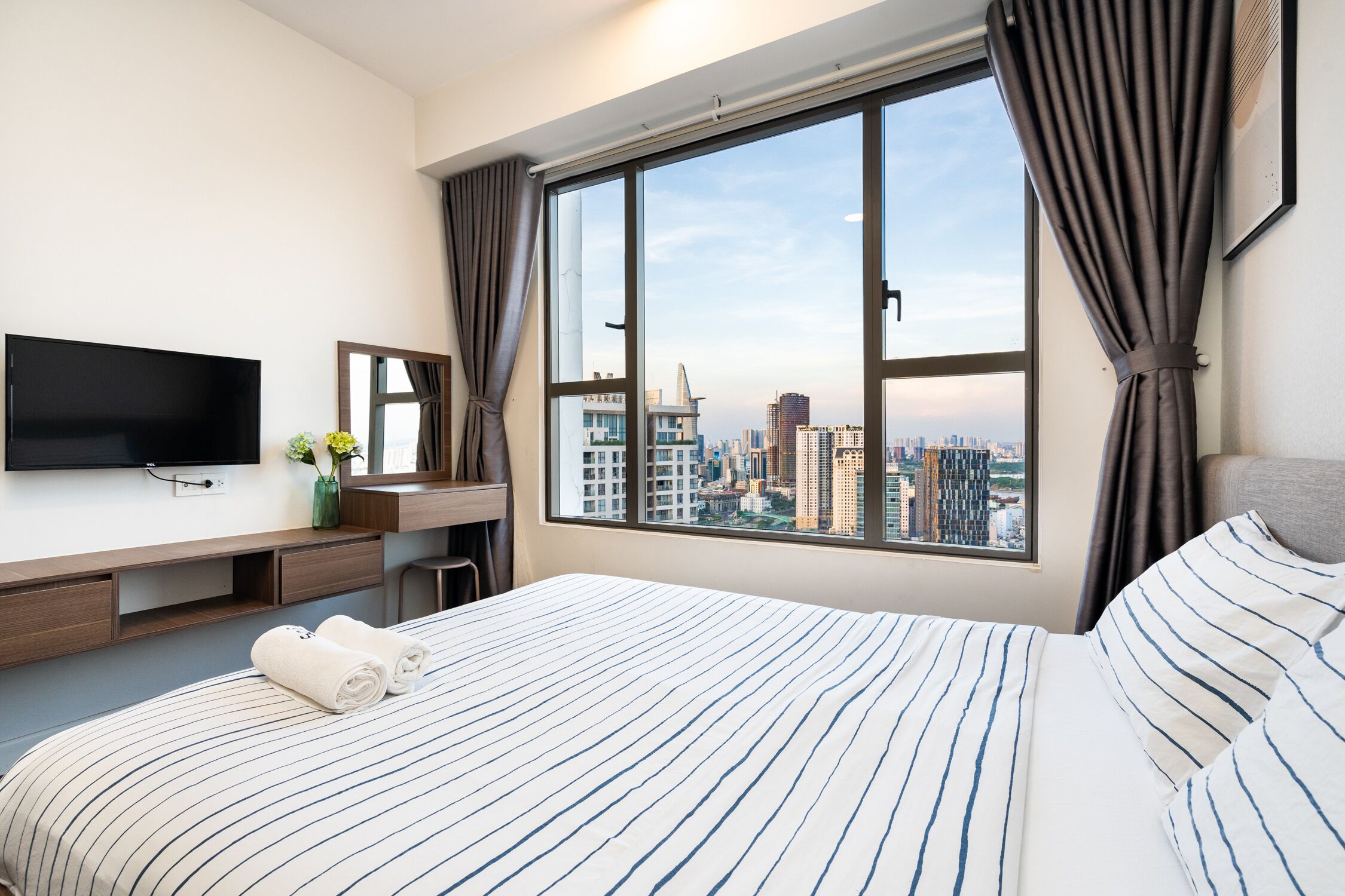 A hotel room with a large bed, a flat-screen TV, a desk with a mirror, and a view of the cityscape through a large window.