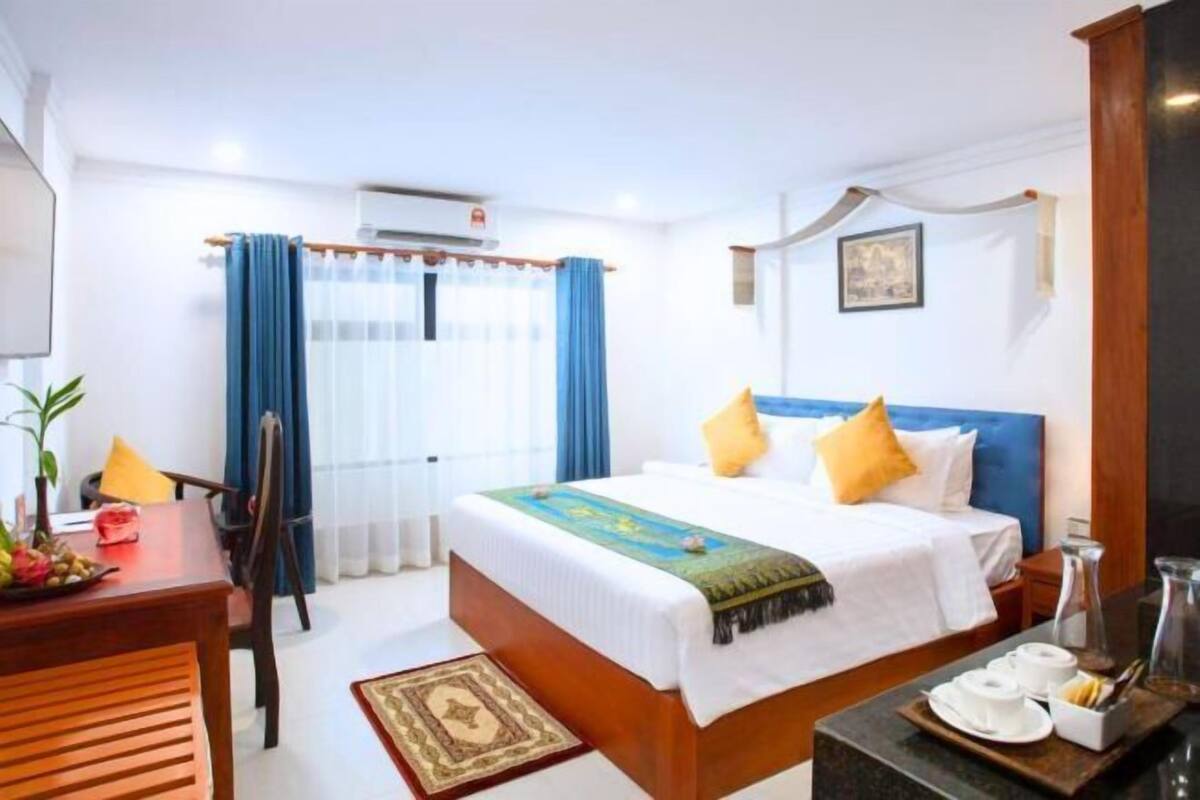 A hotel room with a bed, a desk, a chair, a window with curtains, a wall-mounted air conditioner, and a framed picture.