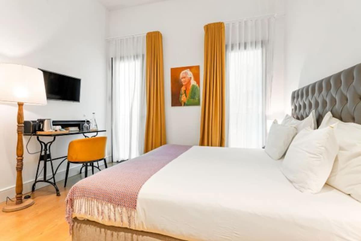 A hotel room with a bed, a desk, a chair, a lamp, and a painting on the wall.