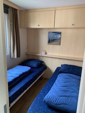 A small, compact room with two bunk beds, a window with curtains, a wall-mounted light, and a painting of a beach scene.