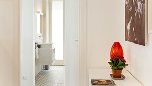 Un baño moderno con un lavabo blanco, un espejo y un jarrón rojo con flores sobre una mesita blanca.