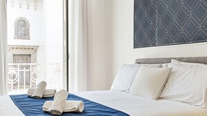 Habitación de hotel con una cama grande, ropa de cama blanca, colcha azul y vista a un edificio a través de una ventana.