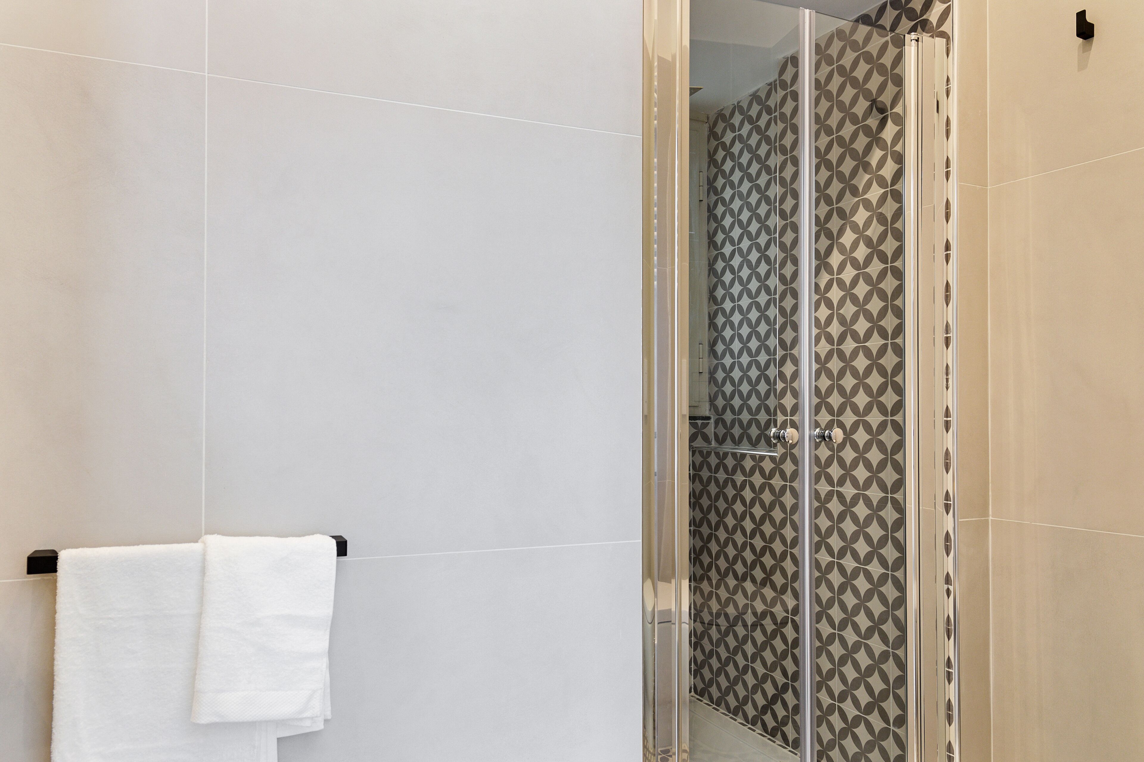 A modern bathroom with a glass shower enclosure, a white towel hanging on a black towel rack, and patterned tiles on the walls.