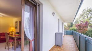 A balcony with a tiled floor, a sliding glass door, a dining area with a table and chairs, and a view of trees and a building in the background. - Buttrio Appartamento nel Verde (Buttrio)