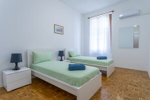 Une chambre avec deux lits, une table de chevet avec une lampe, une fenêtre avec des rideaux et un appareil de climatisation.