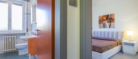 A bedroom with a bed, a bedside table, a nightstand, a window, and a bathroom with a toilet and sink.
