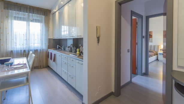 A modern kitchen with white cabinets, a dining table, and a kitchen island. There is a hallway leading to a bedroom with a bed and a nightstand.