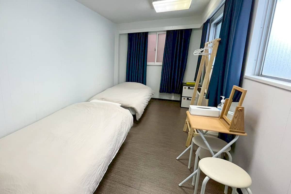 A small, clean room with two single beds, a wooden desk with a chair, a mirror, and a window with curtains.