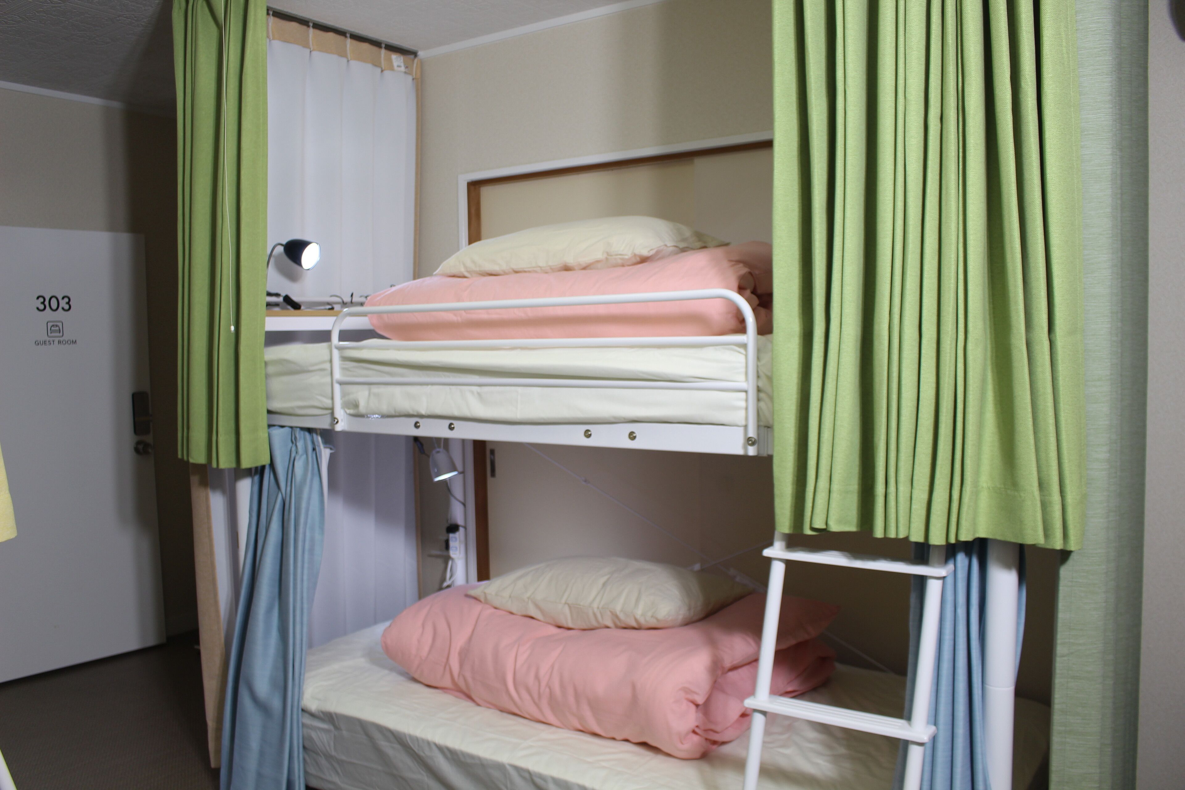 A bunk bed room with a green curtain, a white door with a number, and a small desk with a lamp.
