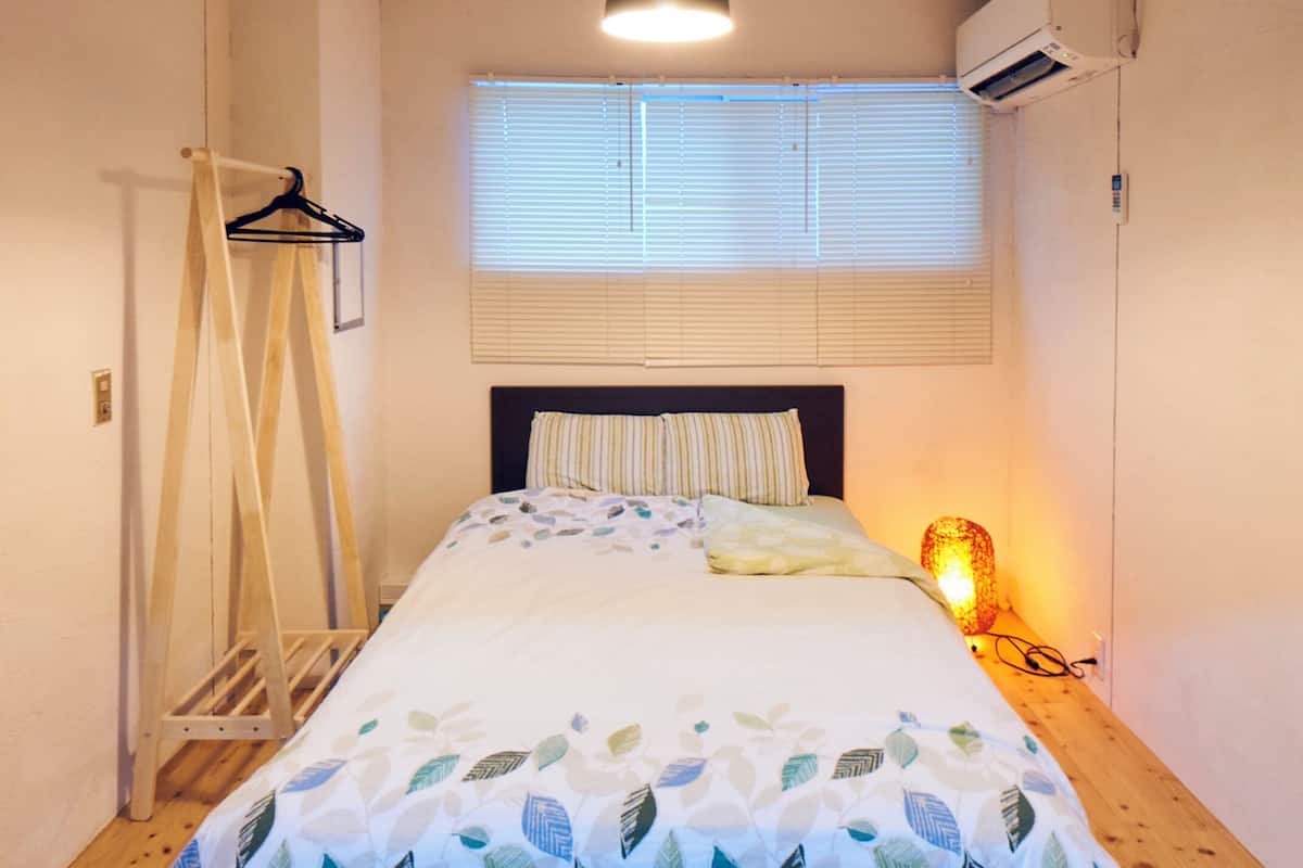 A small, neatly arranged bedroom with a bed, a hanging clothes rack, a window with blinds, and a bedside lamp.