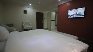 Une chambre d’hôtel avec un grand lit, un fauteuil blanc, un téléviseur fixé au mur et une porte donnant sur une autre pièce.