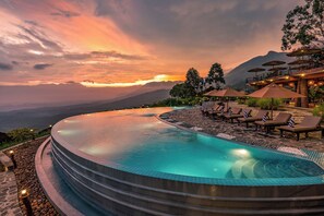 Una piscina infinita con sillas de descanso y sombrillas, rodeada por un sendero de piedra y con vistas a un paisaje montañoso.