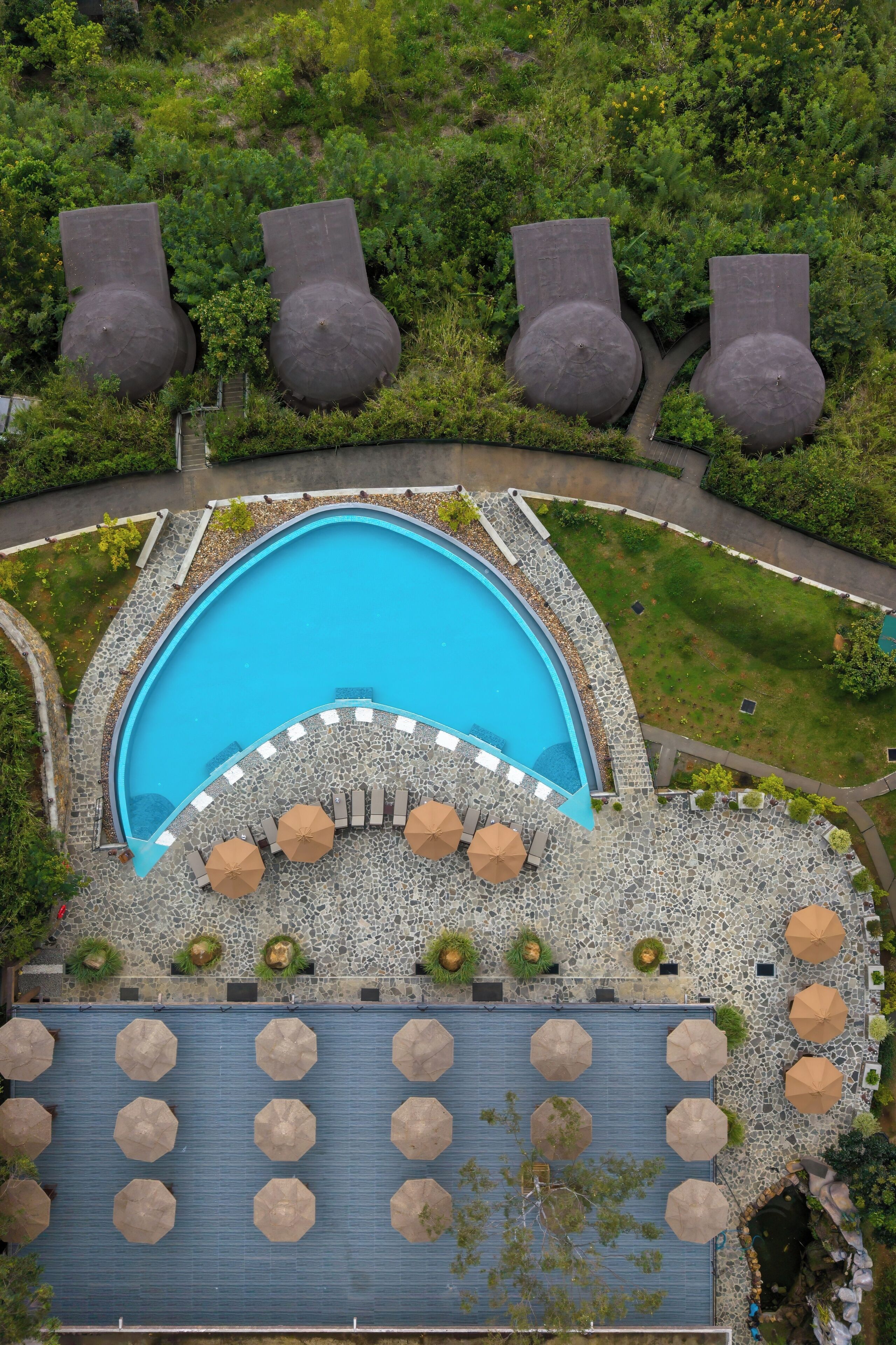 An aerial view of a resort with a swimming pool, lounge area, and surrounding greenery.