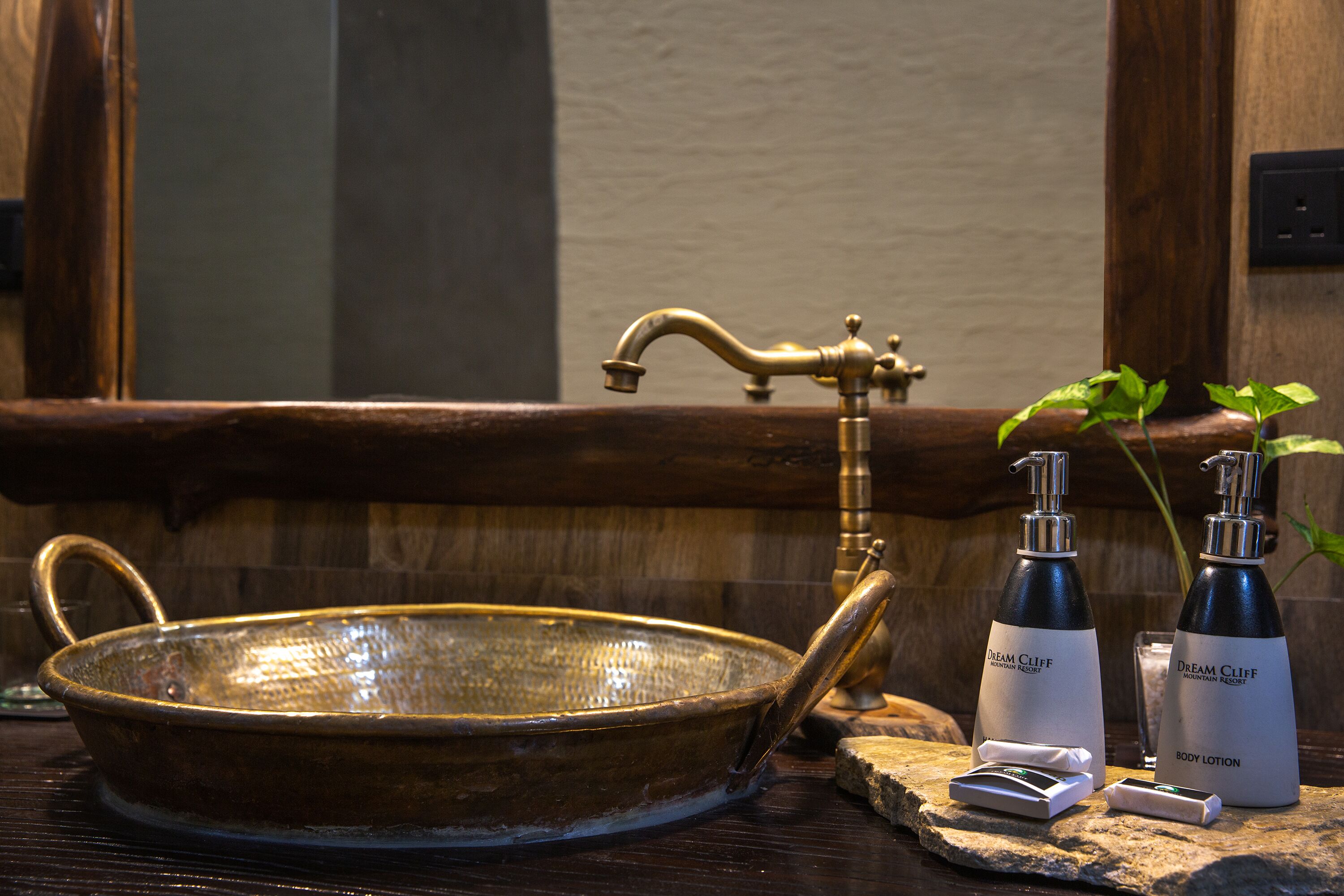 A vintage brass sink with a wooden basin, a classic faucet, and a mirror above it. Two bottles of Dream Cliff body lotion are placed on a stone ledge beside the sink.