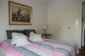 Ein Schlafzimmer mit Bett, Kissen, einem Nachttisch mit Lampe, einer Tür und einem gerahmten Bild an der Wand.