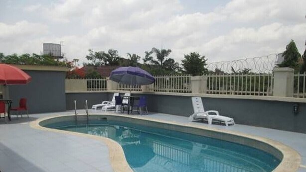 A swimming pool with a curved edge, surrounded by a tiled deck. There are lounge chairs and a table with umbrellas. - Casa Estrella Hotel (Abuja)