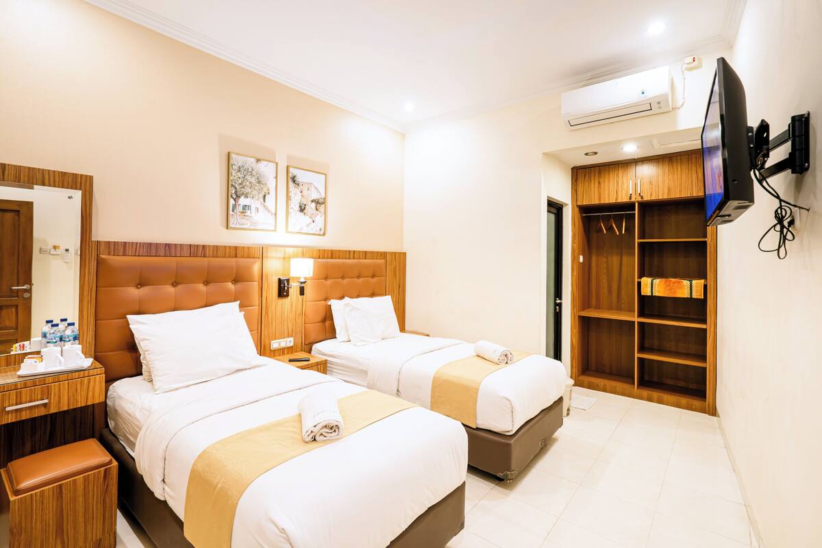 A hotel room with two beds, a wooden wardrobe, a TV mounted on the wall, and an air conditioning unit.